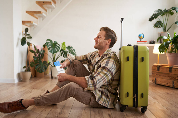 Портрет красивого молодого человека, туриста, сидящего с чемоданом, держащего в руках паспорт и авиабилеты, отправляющегося в отпуск. - Фото, изображение