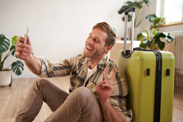 Портрет красивого счастливого парня, турист делает селфи с чемоданом, пользуется смартфоном. Туризм и отдых - Фото, изображение