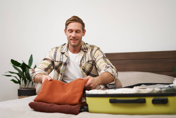 Portrait of handsome young man, tourist unpacking clothes in hotel room, staying in hostel on vacation, travelling, sitting with luggage on bed. - Photo, Image