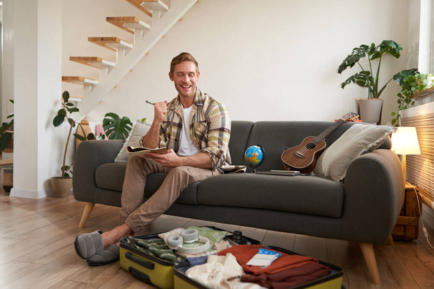 Uomo felice con penna e quaderno, elenco di controllo degli oggetti e valigia per il viaggio, andare in vacanza, prepararsi per le vacanze estive, seduto in soggiorno. - Foto, immagini