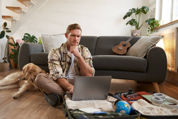Kuva nuoresta miehestä, turisti menossa lomalle, istuu koiran kanssa olohuoneessa, katselee kannettavaa tietokonetta, sisäänkirjautuminen lennolla tietokoneella. Matkailu ja matkustaminen - Valokuva, kuva