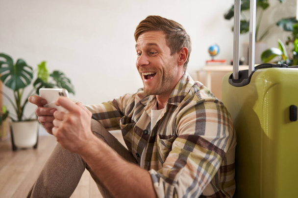 Улыбающийся мужчина с мобильным телефоном, сидит рядом с чемоданом, смеясь, глядя на экран смартфона. Концепция путешествия - Фото, изображение
