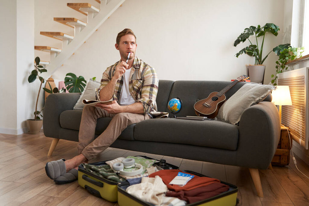 Portrait de jeune homme faisant ses bagages pour un voyage, partant en vacances, mettant des vêtements dans la valise, se préparant pour les vacances. - Photo, image