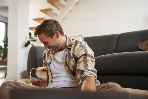 Joyeux jeune homme souriant dans le salon, assis sur le sol, embrassant son animal de compagnie, jouant avec le chien. Concept des personnes et des animaux - Photo, image