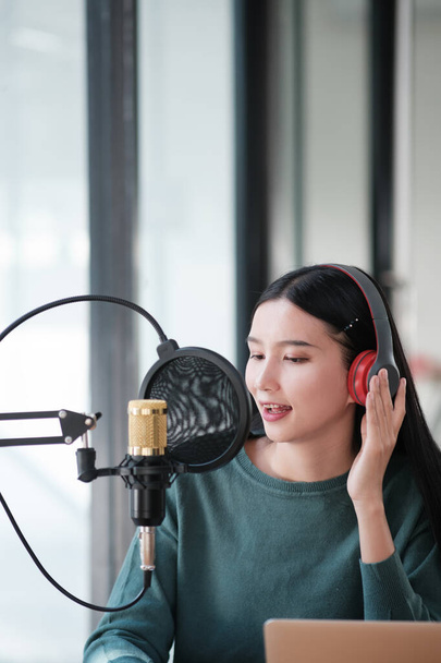 Uma mulher está gravando uma música em um estúdio. Ela está usando fones de ouvido e está sorrindo. O estúdio está equipado com um microfone e um laptop - Foto, Imagem