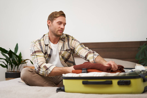 Портрет молодого человека, упаковывающего одежду в чемодан, сидящего на кровати с багажом, отправляющегося в отпуск, готовящегося к путешествию. Концепция образа жизни и туризма - Фото, изображение