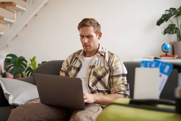 Porträt eines ernsten und konzentrierten Mannes, sitzt mit Laptop im Wohnzimmer, hat zwei Flugtickets, plant Urlaub, geht online. - Foto, Bild