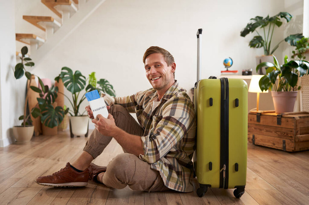 Портрет модели мужчины с чемоданом, сидящего на полу, держащего авиабилеты и паспорт, выезжающего за границу, ожидающего такси до аэропорта. - Фото, изображение
