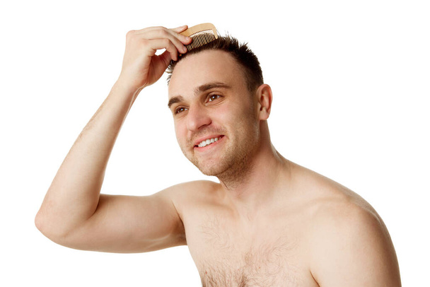 Junger attraktiver Mann ohne Hemd kämmt seine brünetten Haare und schaut vor weißem Studiohintergrund weg. Konzept der natürlichen Schönheit, Wellness-Behandlungen, Maskulinum, Kosmetologie. - Foto, Bild
