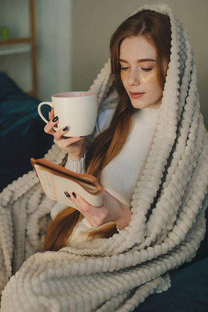 Νεαρή κοκκινομάλλα που κάθεται στον καναπέ καλυμμένη με κουβέρτα, πίνει καφέ και διαβάζει ένα βιβλίο. Κάνω διάλειμμα για καφέ.. - Φωτογραφία, εικόνα