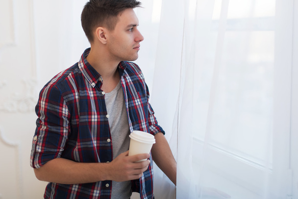 Przystojny facet uśmiechający się człowiek w plaid shirt patrząc przez okno z filiżanka kawa koncepcja rano czeka marzyć - Zdjęcie, obraz