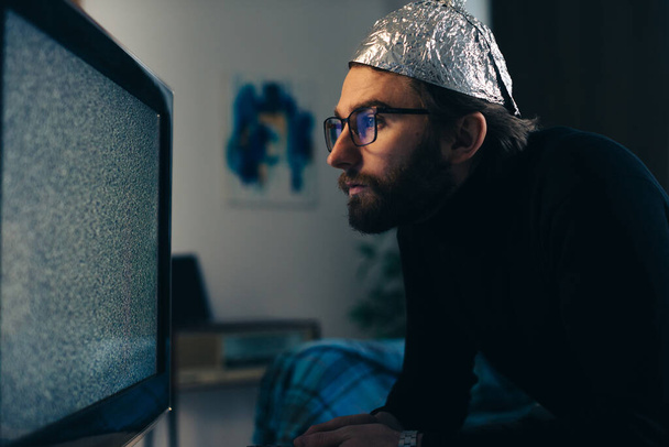 Teoria spiskowa na ekranie: Człowiek w czapce z folii aluminiowej i koc obserwujący zakłócenia telewizyjne - Zdjęcie, obraz