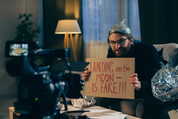 Egy összeesküvés-elmélet elmélet készítője áltudományos videókat készít a kamerán. Egy férfi alufólia kalapban és egy felirat a kezében ül a kanapén a kamera előtt.. - Fotó, kép