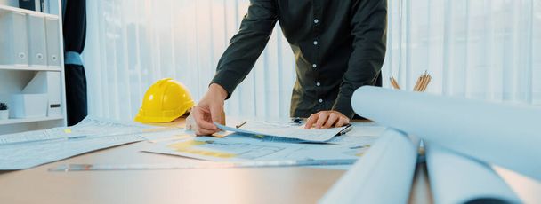 Een portret van professionele mannelijke ingenieur toont foutpunt in blauwdruk op rommelige tafel met architectonisch plan en gele helm geplaatst op modern kantoor. Een close-up. Focus op de hand. Afbakening. - Foto, afbeelding