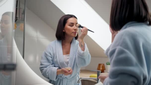 Usmívající se mladá žena nanáší prášek štětcem na make-up v koupelně. Krásná šťastná žena v modrém županu pomocí nové kosmetiky při přípravě doma. Koncept denního make-upu - Záběry, video