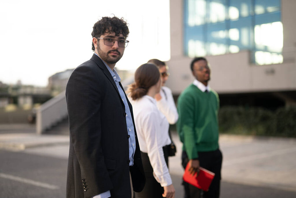 Ένας νεαρός επιχειρηματίας με γυαλιά στέκεται με αυτοπεποίθηση μπροστά, καθώς οι συνάδελφοι συζητούν πίσω του, σε ένα αστικό περιβάλλον.. - Φωτογραφία, εικόνα