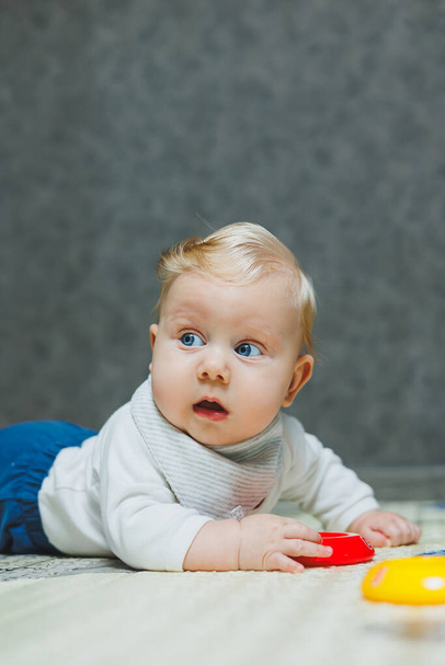 4ヶ月の少年が敷物の上に床に横たわり,おもちゃで遊んでいる. その子は頭を抱えている. 赤ちゃんは床にクロールすることを学ぶ - 写真・画像