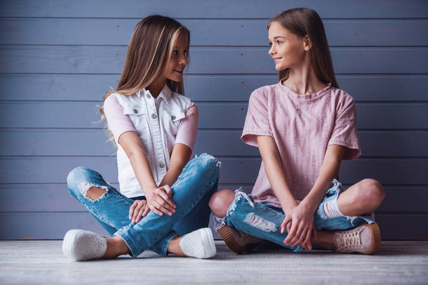 Duas adolescentes atraentes em roupas casuais olhando umas para as outras e sorrindo enquanto se sentam no chão contra um fundo de parede cinza - Foto, Imagem