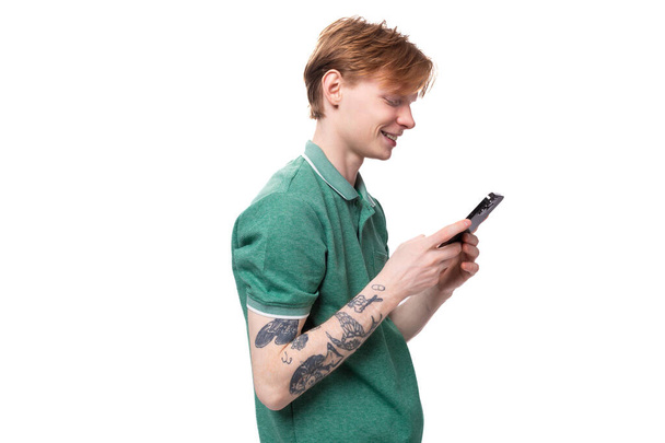 Ένας νεαρός Ευρωπαίος με κόκκινα μαλλιά και τατουάζ στο μπράτσο του γράφει ένα μήνυμα στο τηλέφωνο.. - Φωτογραφία, εικόνα