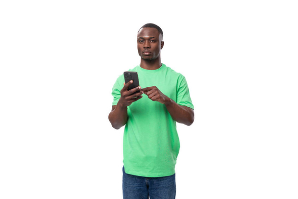 mladý Američan chlap oblečený v mincovní tričko používá telefon na bílém pozadí s kopírovacím prostorem. - Fotografie, Obrázek