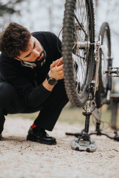 Een zakenman neemt een pauze van zijn werk om zich te concentreren op het repareren van het wiel van zijn fiets op een onverharde weg buiten. - Foto, afbeelding