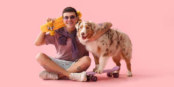 Крутий молодий чоловік і австралійська вівчарка з скейтбордами на рожевому фоні - Фото, зображення