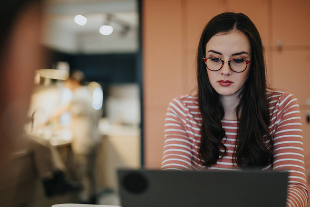 Mujer joven concentrada con gafas que trabajan en el ordenador portátil en la cafetería moderna. - Foto, imagen
