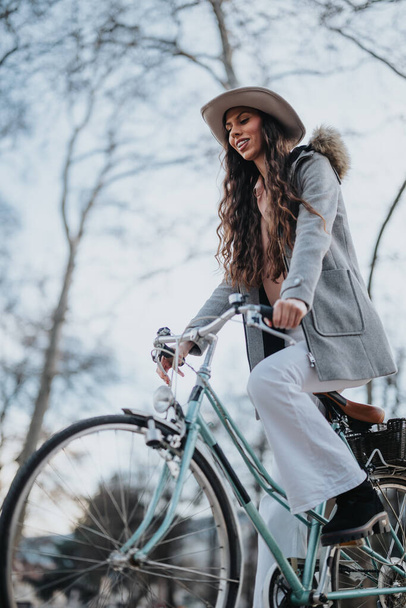 Geniş kenarlı şapkalı modern bir iş kadını, sakin bir park ortamında bisikletini sürüyor.. - Fotoğraf, Görsel