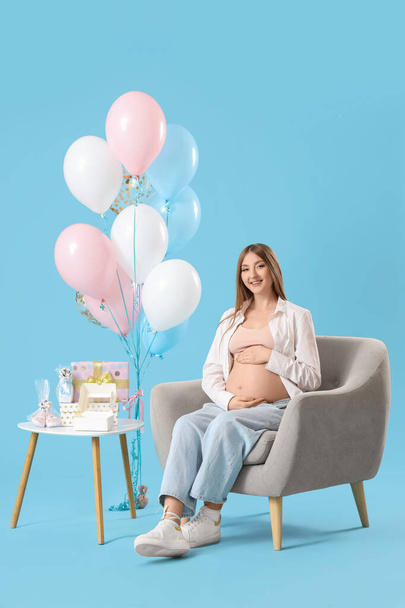 Νεαρή έγκυος γυναίκα με δώρα ντους μωρών και μπαλόνια κάθεται στην πολυθρόνα σε μπλε φόντο - Φωτογραφία, εικόνα