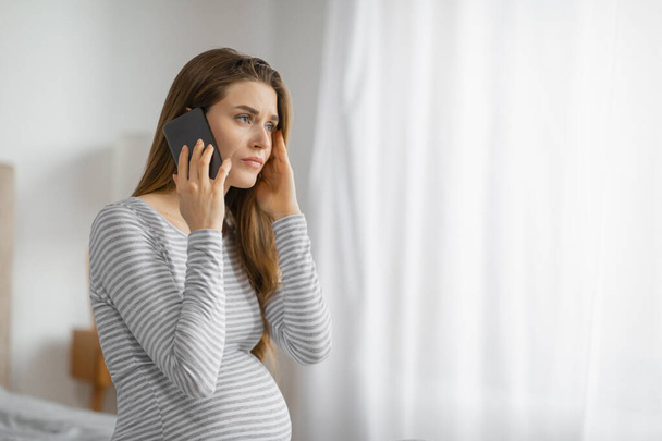 Verwachte moeder aan de telefoon met een bezorgde uitdrukking, staande door een raam - Foto, afbeelding