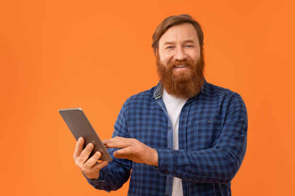 Рудий бородатий бізнесмен випадково використовує цифровий планшетний комп'ютер, посміхаючись камері, стоячи на помаранчевому фоні в студії, портрет чоловіка веб-серфінг на гаджеті - Фото, зображення