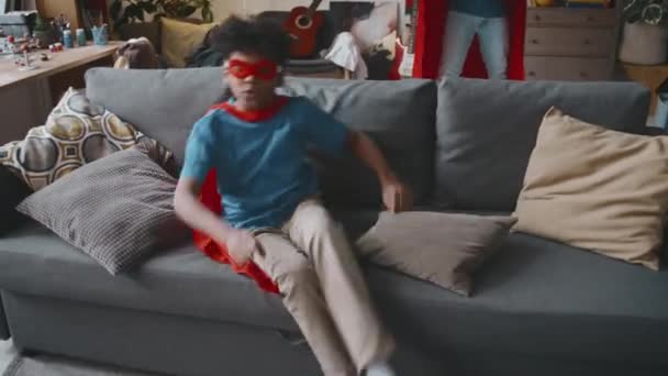 Захоплена афроамериканська дитина в супергеройському костюмі ковзає на дивані, а потім біжить до батька, піднімаючи його і кружляючи навколо з простягнутими руками, граючи разом у вітальні - Кадри, відео