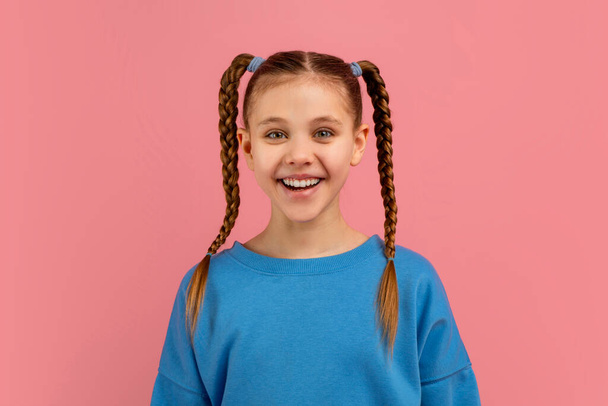 Uzun örgülü saçları olan neşeli bir genç kız pembe bir fona gülümsüyor. - Fotoğraf, Görsel