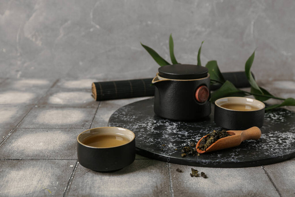 Чайник з чашками, совок сухого чаю і листя на плитковому столі на сірому гранжевому фоні - Фото, зображення