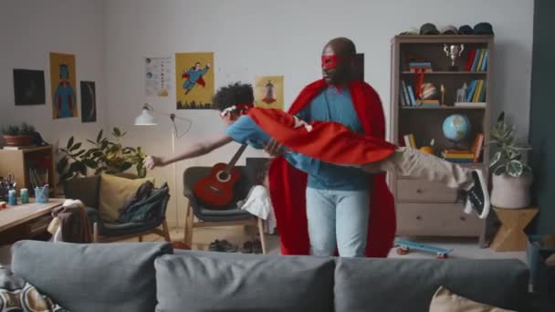Czarny tata trzyma i kręci się wokół małego syna w kostiumie superbohatera podczas zabawy razem w dzień w domu - Materiał filmowy, wideo