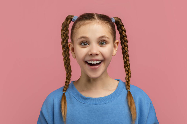 Una giovane ragazza gioiosa con le trecce intrecciate e gli occhi larghi che indossano un top blu, impostato su un solido sfondo rosa - Foto, immagini