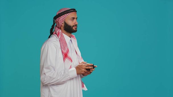 Hombre moderno jugando juegos con controlador, divertirse con la competencia de videojuegos sobre fondo de estudio azul. Jugador islámico en ropa árabe tradicional compite en el torneo. - Foto, Imagen