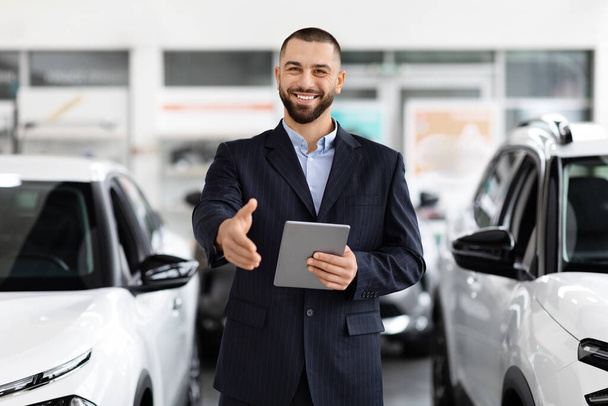 Un vendeur de voiture confiant dans un costume tient une tablette numérique dans une salle d'exposition de voiture, la main tendue et souriant, peut-être prêt à aider - Photo, image