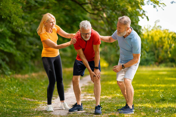 Concerned senior couple helping their male friend recover from injury during outdoor training, elderly man suffering muscle cramp or knee trauma after jogging in green park, free space - Photo, Image