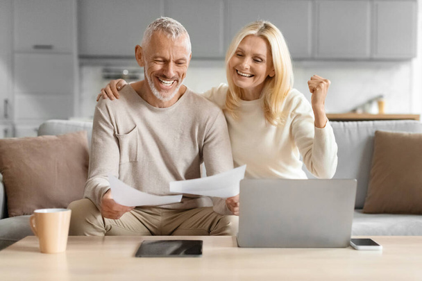 Radosna para seniorów świętująca dobre wieści z papierkową robotą w ręku, siedząca przy laptopie w domu - Zdjęcie, obraz