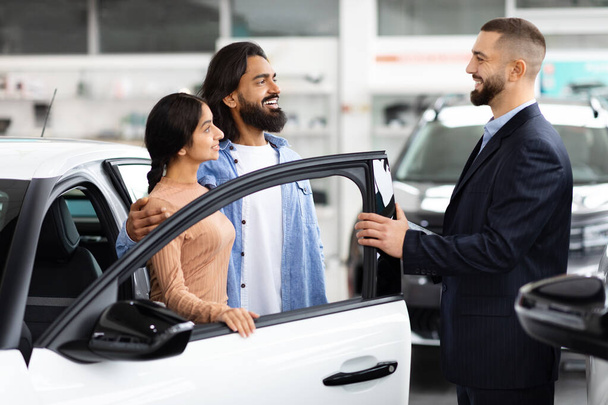 Hymyilevä myyntimies avaa uuden ajoneuvon oven asiakkaille intialainen pari auton näyttelytilassa, joka tarjoaa näkymän sisälle - Valokuva, kuva