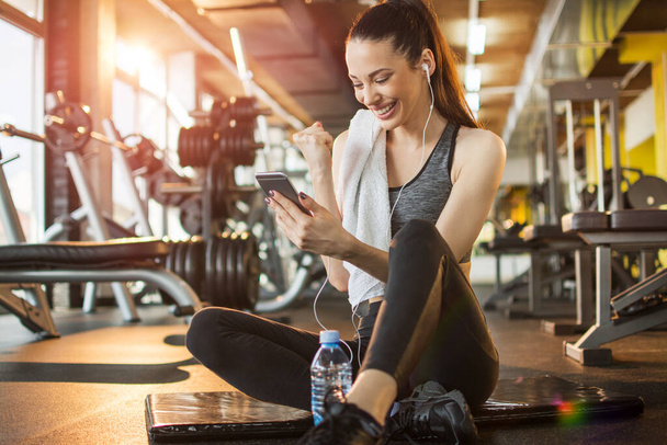 Euforia młoda kobieta fit oglądanie jej smartfon podczas siedzenia na macie do ćwiczeń w siłowni. - Zdjęcie, obraz