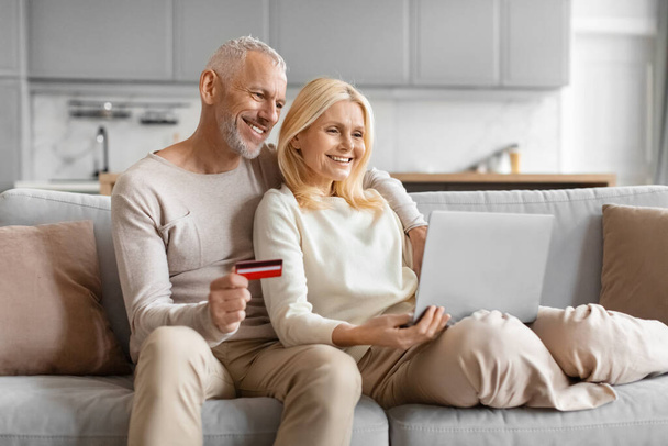 Улыбающаяся взрослая пара с помощью ноутбука и кредитной карты делает онлайн-покупки, сидя на диване - Фото, изображение