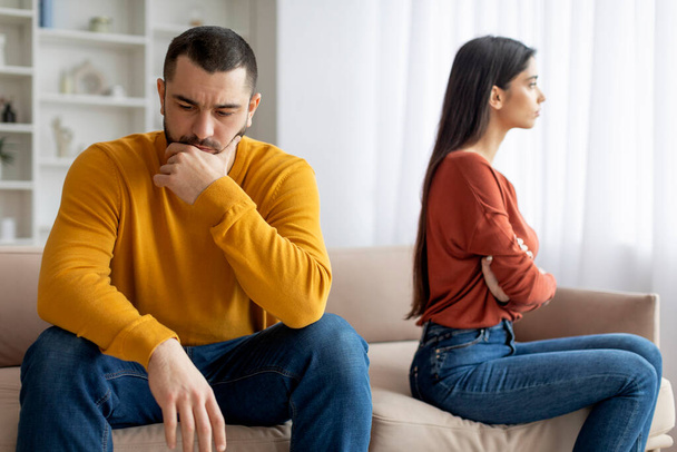 An adult male and female sitting separately on a couch exhibiting signs of a conflict or disagreement, appearing upset and contemplative - Photo, Image