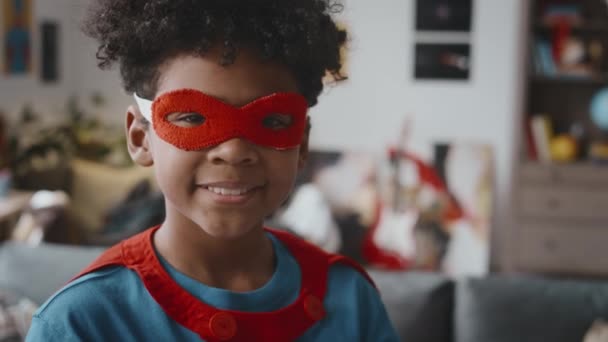 Ritratto di un piccolo ragazzo afroamericano vestito da supereroe che guarda la macchina fotografica e sorride mentre gioca a casa - Filmati, video