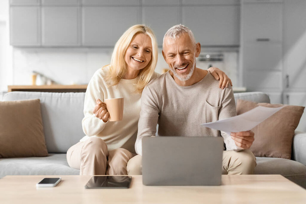 Mature couple excitedly reading a document with coffee in hand, sitting beside a laptop at home - Photo, Image
