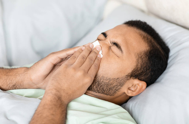 Ungesunder junger schwarzer Mann liegt zu Hause im Bett, niest, leidet unter Erkältung oder Grippe, Nahaufnahme - Foto, Bild