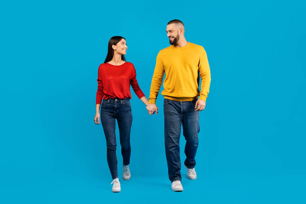 A man and woman are walking hand in hand against blue studio background, showing affection and unity in their relationship, romantic young couple having fun together, copy space - Photo, Image