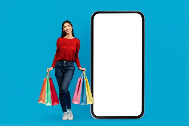 Женщина, стоящая с сумками в руке рядом с большим пустым телефоном, кажется занятой и занимающейся шоппингом, макет - Фото, изображение
