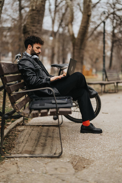 Εστιασμένη αρσενικό επιχειρηματία χρησιμοποιεί ένα φορητό υπολογιστή, ενώ κάθεται σε ένα παγκάκι πάρκο, παραδειγματική κινητικότητα και σύγχρονο στυλ εργασίας. - Φωτογραφία, εικόνα